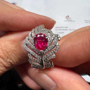 May include: A silver-toned ring featuring a large, pear-shaped, vibrant red gemstone at the centre. The band and setting are designed in a detailed feather motif, encrusted with numerous small, clear, sparkling white stones throughout the metal.