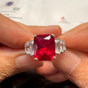 May include: A silver-toned ring featuring a large, rectangular, vibrant red center stone held by four prongs. The band is accented on each side by two smaller, clear, baguette-cut stones, creating a symmetrical design with a polished metallic finish.