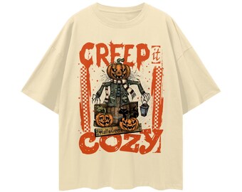 May include: Cream-colored t-shirt with a Halloween-themed graphic. The design features a scarecrow with a pumpkin head, the words "CREEP it COZY", and two jack-o'-lanterns. The shirt has short sleeves and a relaxed fit.