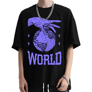 Puede incluir: Camiseta negra con un gráfico morado de una mano tocando un globo terráqueo, con la palabra "WORLD" en letras moradas grandes debajo. El diseño incluye cuatro formas de estrella moradas. La camiseta tiene mangas cortas.