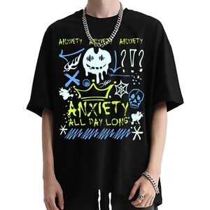Puede incluir: Camiseta negra con un diseño gráfico que presenta la palabra "ANXIETY" en amarillo y blanco, junto con varios garabatos en azul y blanco. El diseño incluye una corona, una calavera y signos de interrogación.