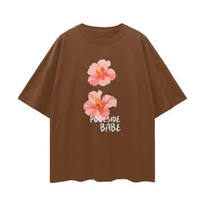Peut inclure: Un t-shirt marron avec deux fleurs d'hibiscus roses et oranges. Le texte "Poolside Babe" est imprimé en blanc en dessous des fleurs. Le t-shirt a des manches courtes et un col rond.