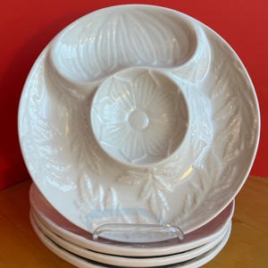 May include: A stack of white ceramic serving dishes. Each dish has a central well and a curved section, with a floral and leaf pattern. The dishes are stacked on a clear plastic stand, set against a red background.