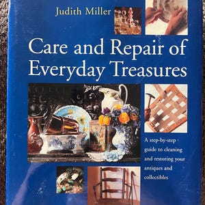 Peut inclure: Un livre relié bleu intitulé Care and Repair of Everyday Treasures par Judith Miller. La couverture présente plusieurs photographies d'antiquités, dont de la verrerie, une chaise et une personne utilisant un marteau sur un siège tressé, avec le texte Reader's Digest.