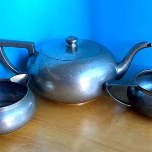 May include: A vintage silver tea set comprising a teapot with lid and two small serving bowls. The metal set has a classic design. The background is a solid blue. The set is made of metal.