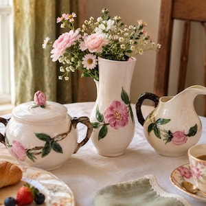 Puede incluir: Un juego de té de cerámica color crema con diseños florales rosas, que incluye una azucarera, una jarra de crema y un jarrón. La azucarera tiene una tapa en forma de rosa. Una taza de café y un cruasán también están sobre la mesa.
