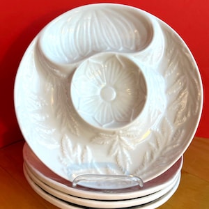 May include: A stack of white ceramic serving dishes with a floral and leaf design. Each dish has a central dip bowl and a curved section. The dishes are stacked on a clear plastic stand, with a red background and a wooden surface.