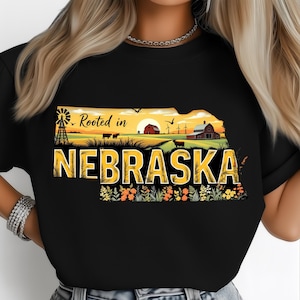 Op de afbeelding: Zwart T-shirt met een Nebraska-vormig ontwerp met een landelijk landschap. Het ontwerp bevat een windmolen, boerderijgebouwen, koeien en de tekst "Rooted in Nebraska" en "NEBRASKA" in het geel.