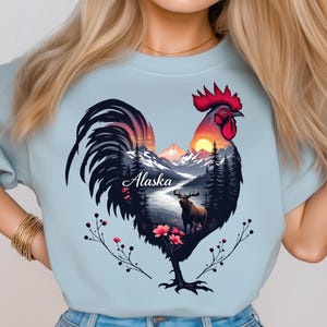 Puede incluir: Camiseta azul claro con un diseño de gallo. El cuerpo del gallo contiene un paisaje de Alaska con montañas, árboles y un alce. El gallo tiene una cresta y barbillas rojas, y una cola negra. La palabra "Alaska" está escrita en cursiva.