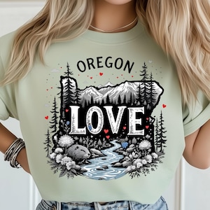 Puede incluir: Camiseta verde claro con un gráfico del estado de Oregón y la palabra "LOVE" en blanco. El diseño incluye un río, montañas, árboles y un castor. La palabra "OREGON" está encima del contorno del estado.