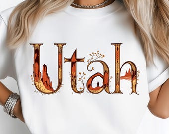 May include: White t-shirt featuring the word "Utah" in a decorative, desert-themed design. The letters are in shades of brown, orange, and yellow, with illustrations of rock formations and a person under an arch. A stylish and comfortable tee.