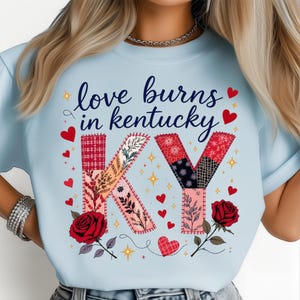 May include: Pale blue t-shirt featuring the phrase "love burns in kentucky" in dark blue script. The letters "KY" are designed with patchwork patterns, surrounded by red hearts, roses, and gold stars. A romantic and stylish design.