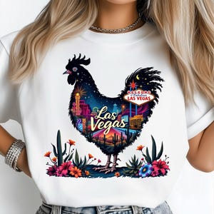 Puede incluir: Camiseta blanca con un diseño de gallo negro con temática de Las Vegas. El cuerpo del gallo está lleno de una colorida ilustración del horizonte de Las Vegas, un paisaje desértico y las palabras "Las Vegas". La camiseta está decorada con flores.