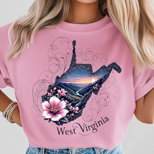 Puede incluir: Camiseta rosa con un gráfico detallado del contorno del estado de Virginia Occidental, lleno de un paisaje de montañas, un río y una puesta de sol. El diseño incluye flores rosas y el texto "West Virginia".