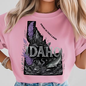 Op de afbeelding: Roze t-shirt met een gedetailleerde afbeelding van de staat Idaho. Het ontwerp bevat een bergketen, rivier, vis en bloemelementen, met het woord "IDAHO" en de tekst "PRIDE IN THIS LAND".