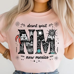 Può includere: T-shirt rosa chiaro con un design grafico con le lettere "NM" riempite di immagini del deserto, tra cui cactus, un sole e edifici. Le parole "desert spirit" e "new mexico" sono stampate anche sulla maglietta.