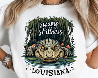 May include: White t-shirt featuring a detailed illustration of an alligator in a swamp setting. The design includes the text "swamp stillness" above the alligator and "LOUISIANA" below, with musical notes. The alligator is in a pond with lily pads and flowers.