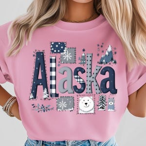 May include: Pink t-shirt with the word "Alaska" in a patchwork design. The letters are decorated with patterns, snowflakes, trees, and a polar bear. The shirt has a crew neck and short sleeves.
