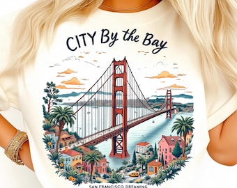 Puede incluir: Camiseta color crema con una colorida ilustración del Golden Gate Bridge en San Francisco. El diseño incluye el texto "CITY By the Bay" y "SAN FRANCISCO DREAMING". Palmeras y edificios rodean el puente.