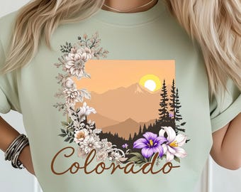 May include: A light green t-shirt featuring a floral design and the word "Colorado" in a brown script. The design includes a mountain landscape with a sunset, trees, and flowers in white, purple, and cream colors.