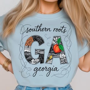 Könnte beinhalten: Hellblaues T-Shirt mit dem Text "southern roots GA georgia". Die Buchstaben "GA" sind mit Illustrationen eines Baumes, Bergen, einem Pfirsich und einem gelben Vogel gefüllt. Das Design ist von einer dekorativen Bordüre umrandet.