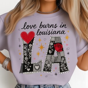 May include: A lilac t-shirt with the words "love burns in louisiana" printed on it. The letters "LA" are decorated with floral and heart designs. A red heart and rose are also featured in the design.