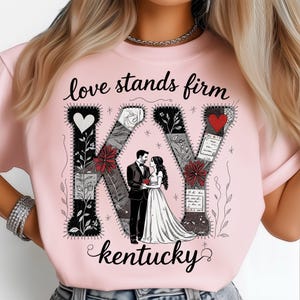 May include: A pale pink t-shirt with the text "love stands firm" and "kentucky" in black script. The design includes a couple in formal wear, with the letters "KY" decorated with floral and heart motifs.