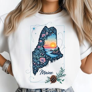 Puede incluir: Camiseta blanca con una ilustración detallada del estado de Maine. El diseño incluye una escena de puesta de sol dentro del contorno del estado, llena de flores y follaje. La palabra "Maine" está escrita en cursiva debajo.