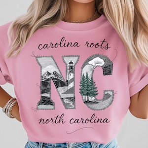 Puede incluir: Camiseta rosa claro con las palabras "carolina roots" y "north carolina" en escritura negra. Las letras "NC" están rellenas con imágenes de montañas, un faro y árboles, con un efecto cosido.