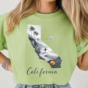 Puede incluir: Sudadera verde claro con un diseño de California. El diseño muestra una escena de playa con palmeras, un surfista y la palabra "California". El gráfico utiliza tonos grises, blancos y naranjas.