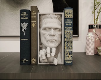 May include: A bookend featuring a sculpted face of Frankenstein's monster emerging from a book. The bookend is made of white plaster and is holding a book with the title "The Jungle Book" by Rudyard Kipling.