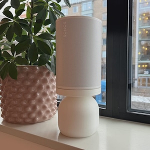 Puede incluir: Un altavoz Sonos blanco con el nombre de la marca en la parte superior, sobre un soporte blanco. A la izquierda del altavoz hay una maceta de cerámica rosa texturizada con una planta verde. El altavoz está cerca de una ventana.