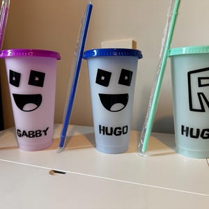 Puede incluir: Tres vasos reutilizables de colores con pajitas y nombres personalizados: Gabby, Hugo y Hugh. Cada vaso presenta un dise&ntilde;o diferente, incluyendo una cara sonriente y una letra R.