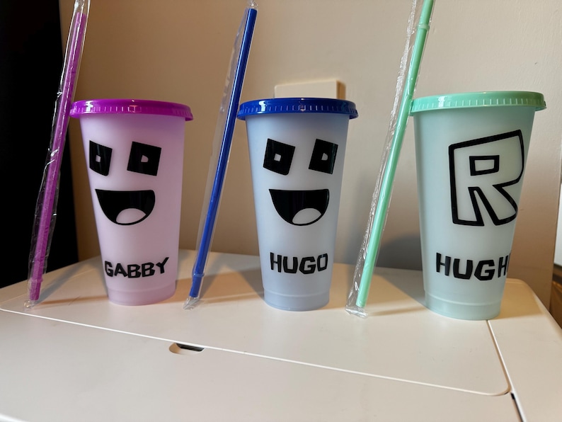 Puede incluir: Tres vasos reutilizables de colores con pajitas y nombres personalizados: Gabby, Hugo y Hugh. Cada vaso presenta un dise&ntilde;o diferente, incluyendo una cara sonriente y una letra R.