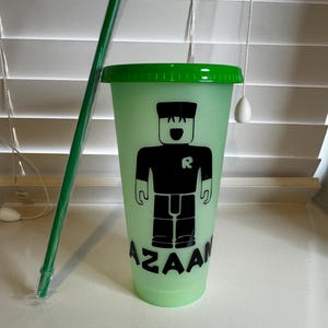 Puede incluir: Un vaso reutilizable verde y negro con una tapa verde y una pajita. El vaso tiene un gr&aacute;fico de un personaje de Roblox con el texto "AZAAN" impreso en la parte inferior.