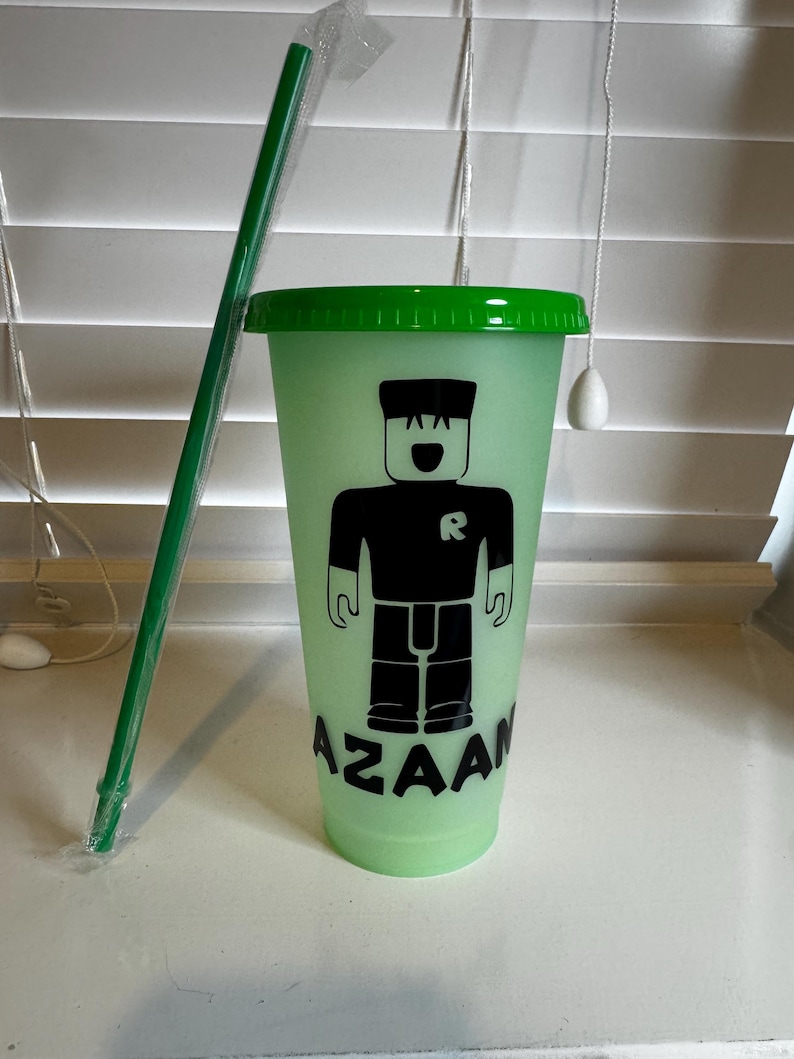 Puede incluir: Un vaso reutilizable verde y negro con una tapa verde y una pajita. El vaso tiene un gr&aacute;fico de un personaje de Roblox con el texto "AZAAN" impreso en la parte inferior.
