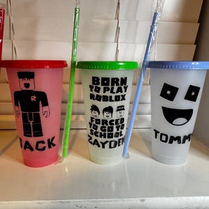 Puede incluir: Tres vasos reutilizables de colores con diferentes dise&ntilde;os y nombres. El vaso rosa tiene un personaje de Roblox y el nombre "Jack". El vaso verde tiene un dise&ntilde;o que dice "Born to Play Roblox Forced to Go to School Zayden". El vaso azul tiene una cara sonriente y el nombre "Tommy".