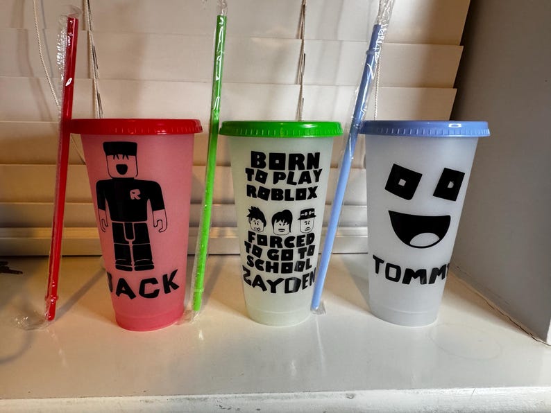 Puede incluir: Tres vasos reutilizables de colores con diferentes dise&ntilde;os y nombres. El vaso rosa tiene un personaje de Roblox y el nombre "Jack". El vaso verde tiene un dise&ntilde;o que dice "Born to Play Roblox Forced to Go to School Zayden". El vaso azul tiene una cara sonriente y el nombre "Tommy".