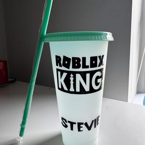 May include: A reusable cup with a green lid and a black design that says "Roblox King with a personalised name". The cup has a straw.