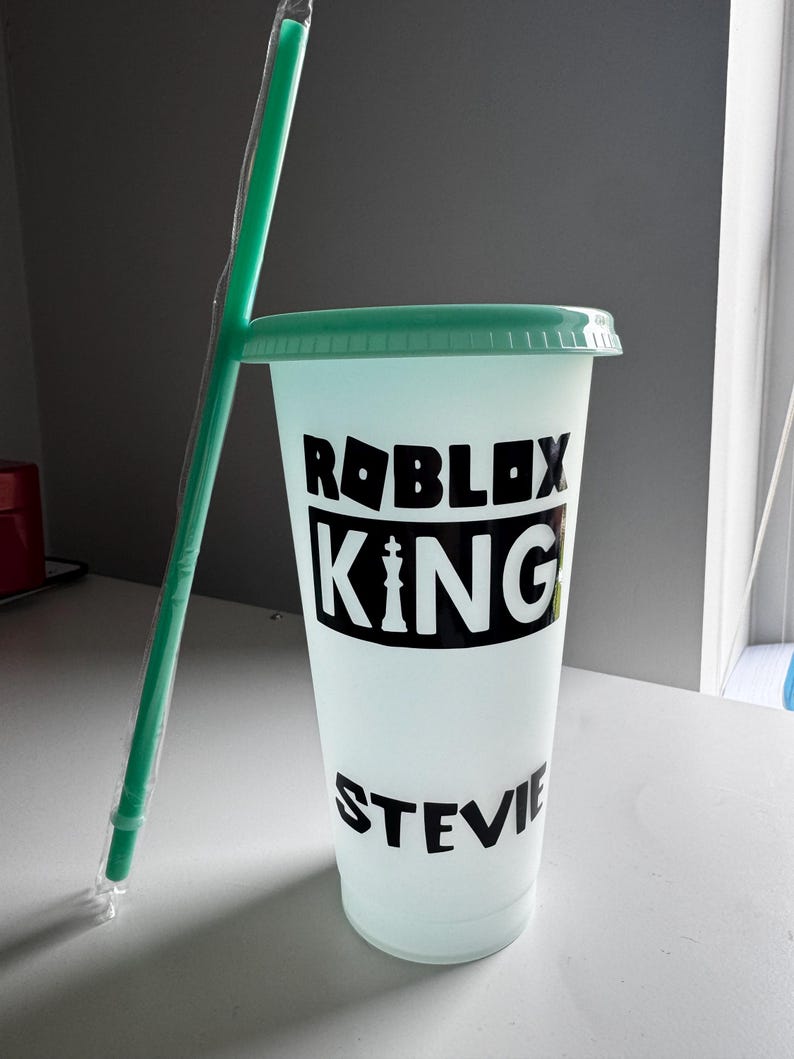 May include: A reusable cup with a green lid and a black design that says "Roblox King with a personalised name". The cup has a straw.