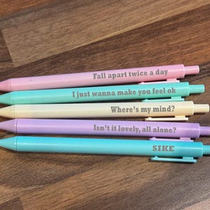 May include: Five pastel-coloured pens with black text. The pens are pink, teal, cream, lavender, and light blue. Each pen has a different phrase: "Fall apart twice a day", "I just wanna make you feel ok", "Where's my mind?", "Isn't it lovely, all alone?", and "SIKE".