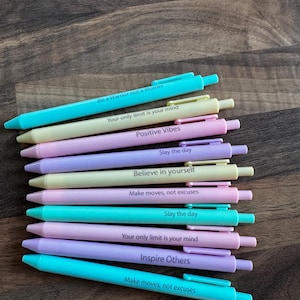 May include: An assortment of pastel-coloured pens, each bearing an inspirational quote. The pens are in hues of blue, yellow, and lilac. Phrases such as "Believe in yourself" and "Slay the day" are visible.