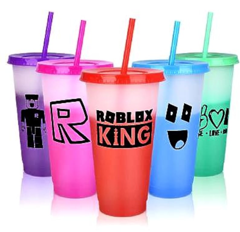 Set of  colourful reusable tumblers with lids and straws. Each tumbler features a different design, including a Roblox King logo, a smiley face, and a peace sign with a heart.