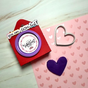 Puede incluir: Una caja roja con asa y el texto "you are -DOH-best!" en la parte delantera. La caja contiene un recipiente de Play-Doh morado con una etiqueta blanca que dice "Happy Valentine's Day". Una hoja de papel rosa con recortes de corazones blancos está en el fondo. Un corazón de Play-Doh morado y un cortador de galletas en forma de corazón blanco están sobre el papel rosa.