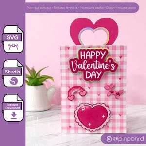 Puede incluir: Una bolsa de regalo de San Valentín con un patrón de cuadros rosas y blancos. La bolsa presenta un asa en forma de corazón y está decorada con un letrero de "Happy Valentine's Day", un arcoíris, un corazón y un perro globo.