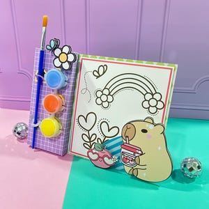 Puede incluir: Un kit de pintura por números con un lindo capibara de dibujos animados bebiendo una bebida con una fresa en la parte superior. El kit incluye una paleta de pintura con tres colores: azul, naranja y amarillo. Se incluye un pincel.