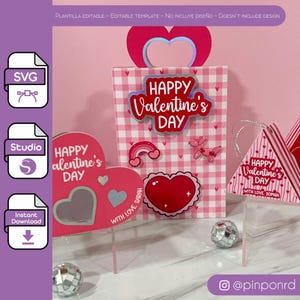 Puede incluir: Un conjunto de regalos temáticos de San Valentín, que incluyen una bolsa de regalo a cuadros rosa y blanco con un asa en forma de corazón y un letrero de "Happy Valentine's Day". También hay tarjetas en forma de corazón.