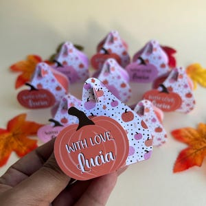 Puede incluir: Un conjunto de pequeñas cajas con forma de calabaza con fondo blanco y lunares rosas. Las cajas están decoradas con una pequeña calabaza y el texto "With love, Lucia".