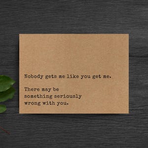 May include: A rectangular tan card with black typewriter-style text that reads: Nobody gets me like you get me. There may be something seriously wrong with you. The card is positioned next to a small green leafy branch on a dark wood surface.