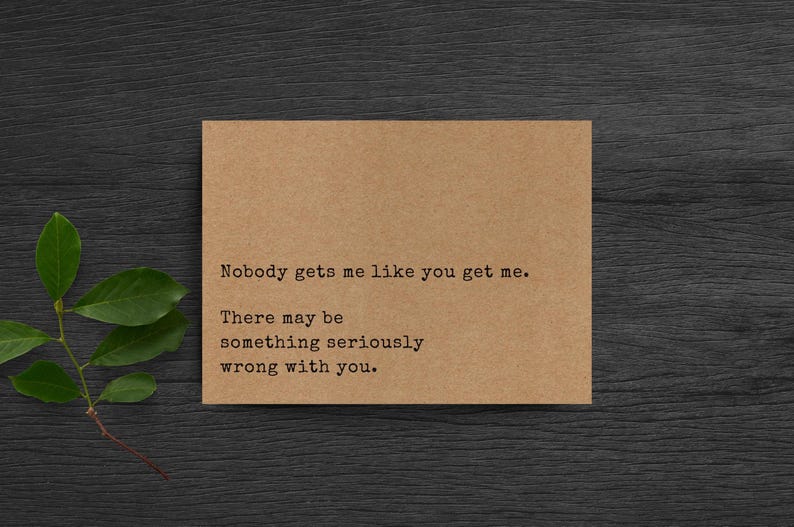 May include: A rectangular tan card with black typewriter-style text that reads: Nobody gets me like you get me. There may be something seriously wrong with you. The card is positioned next to a small green leafy branch on a dark wood surface.